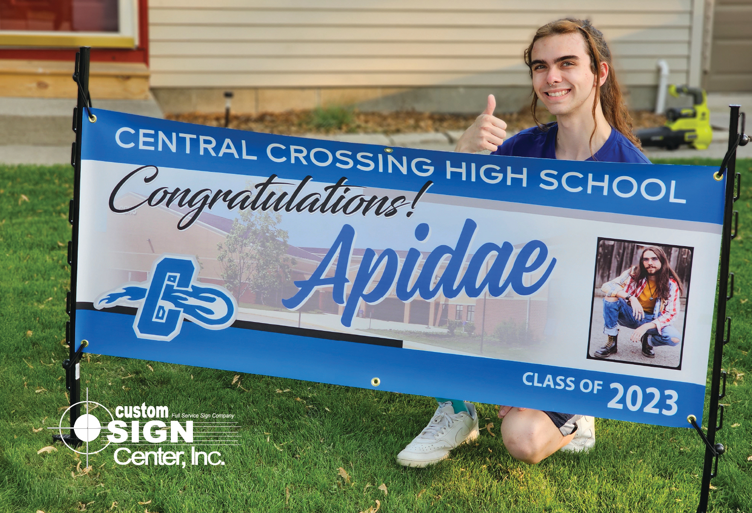 Graduate smiling and giving a thumbs up next to a personalized graduation banner displayed in the yard